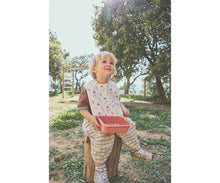 Load image into Gallery viewer, A toddler sitting outdoors on a wooden stool wearing a patterned bib and plaid outfit, holding a pink tray. Trees and a sunny outdoor setting are visible in the background.