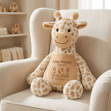 Load image into Gallery viewer, A personalized plush giraffe toy sitting on a cream upholstered armchair in a softly lit nursery. The giraffe has tan and cream spots and a custom tan belly patch with the name 'Liam Stanley Flynn' and birth details.