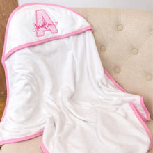 Load image into Gallery viewer, White hooded towel with pink trim and embroidered letter 'A' on a beige cushion.