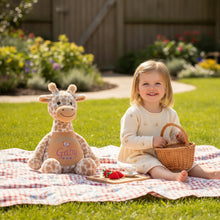 Load image into Gallery viewer, A plush giraffe toy with a beige and brown spotted pattern, featuring a custom embroidered name 'Gaby' and the phrase 'I belong to' on its belly, set with a small child having a picnic in the garden. 